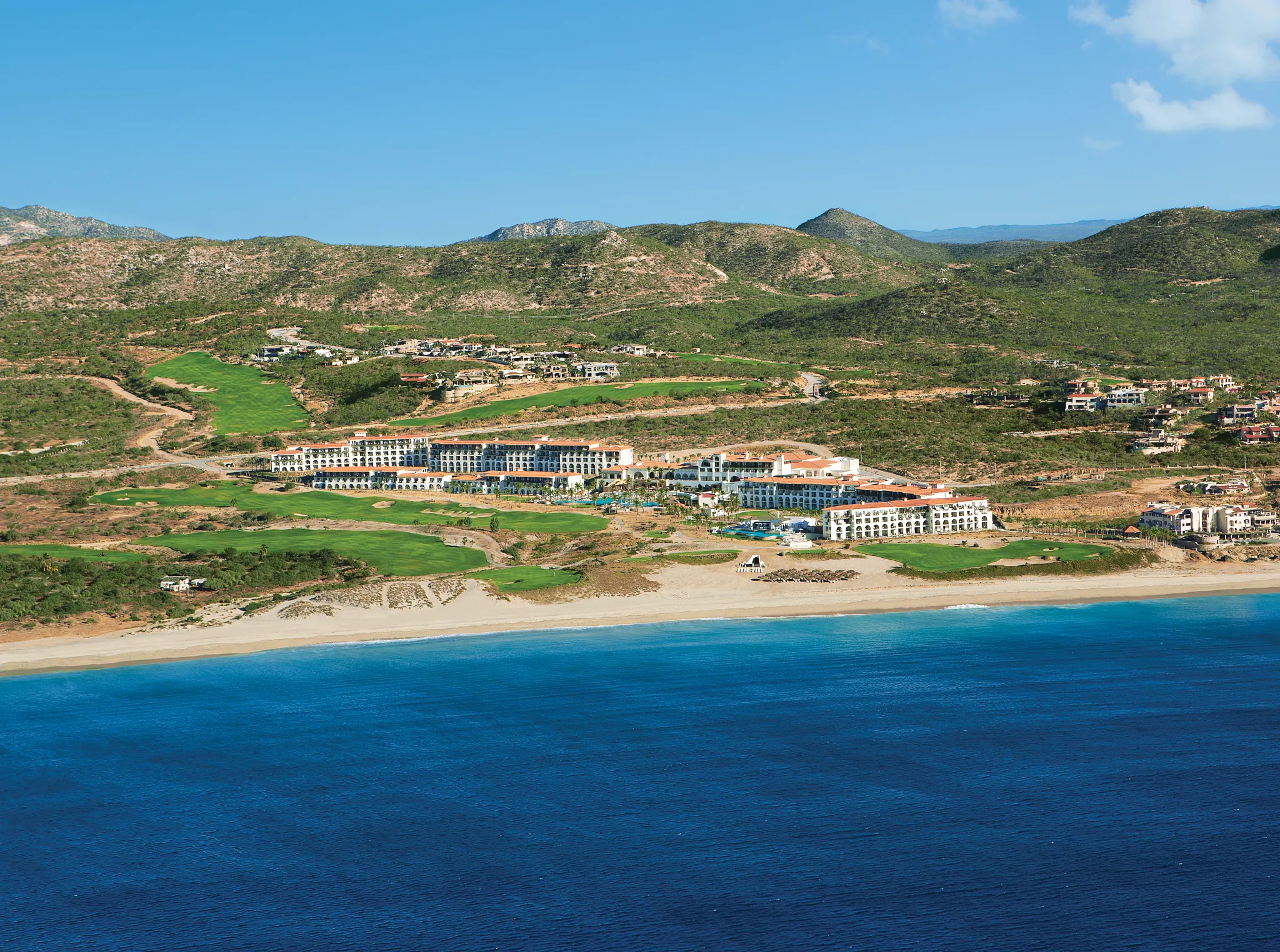 Wide aerial view of Secrets Puerto Los Cabos resort along the beach with mountains and golf course.